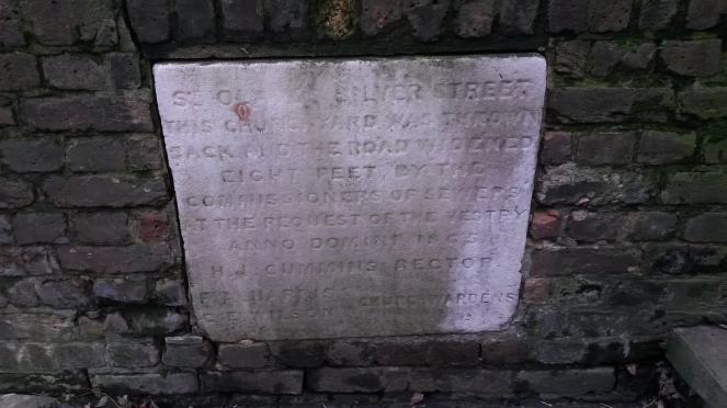 Close up image of the plaque described in the previous paragraph.  It is made of a pale stone, and is set within a wall made of dark-coloured bricks..